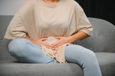 Genç bir kadın evdeki kanepede otururken karın ağrısı çekiyor. Karın ağrısının nedeni adet sancısı, mide iltihabı, mide ülseri, gıda zehirlenmesi, ishal ya da bağırsak iltihabı. Seçici odak.