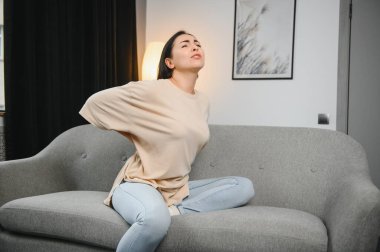 Sırt ağrısı çeken olgun bir kadın oturma odasındaki koltukta oturan mutsuz, yaşlı bir sarışın sırt ağrısı yüzünden rahatsız..