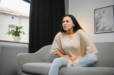 Genç bir kadın evdeki kanepede otururken karın ağrısı çekiyor. Karın ağrısının nedeni adet sancısı, mide iltihabı, mide ülseri, gıda zehirlenmesi, ishal ya da bağırsak iltihabı. Seçici odak.