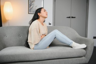 Güzel İspanyol bir kadın, acı dolu bir ifadeyle karnını tutuyor. Adet sancısı çekiyor. Evde üzgün yatıyor, kadın sağlığı konseptinde karnı ağrıyor..