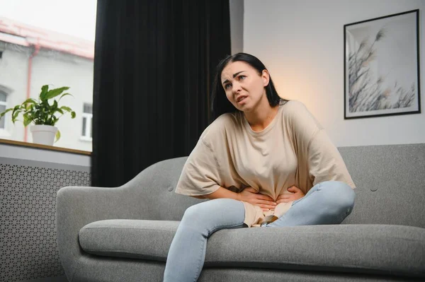 Genç bir kadın evdeki kanepede otururken karın ağrısı çekiyor. Karın ağrısının nedeni adet sancısı, mide iltihabı, mide ülseri, gıda zehirlenmesi, ishal ya da bağırsak iltihabı. Seçici odak.