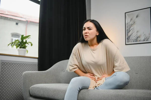 Genç bir kadın evdeki kanepede otururken karın ağrısı çekiyor. Karın ağrısının nedeni adet sancısı, mide iltihabı, mide ülseri, gıda zehirlenmesi, ishal ya da bağırsak iltihabı. Seçici odak.