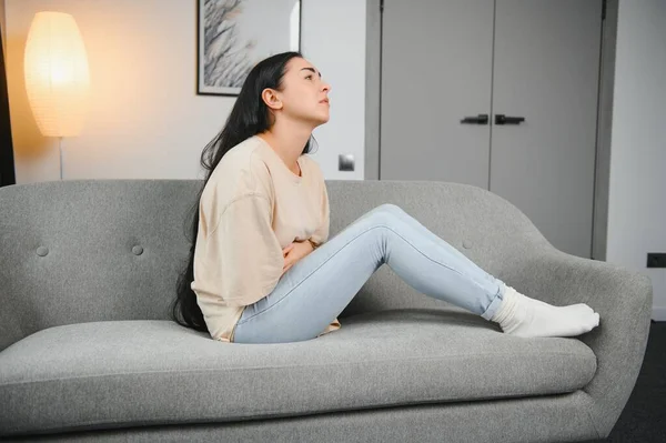 Güzel İspanyol bir kadın, acı dolu bir ifadeyle karnını tutuyor. Adet sancısı çekiyor. Evde üzgün yatıyor, kadın sağlığı konseptinde karnı ağrıyor..