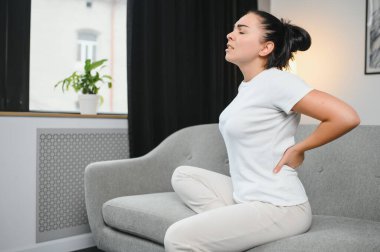 Sırt ağrısı çeken olgun bir kadın oturma odasındaki koltukta oturan mutsuz, yaşlı bir sarışın sırt ağrısı yüzünden rahatsız..