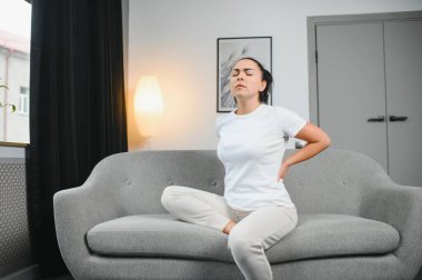 Sırt ağrısı çeken olgun bir kadın oturma odasındaki koltukta oturan mutsuz, yaşlı bir sarışın sırt ağrısı yüzünden rahatsız..