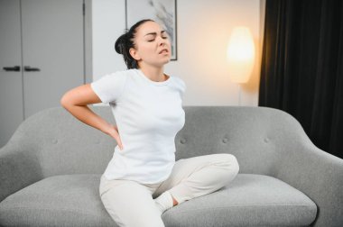 Sırt ağrısı çeken olgun bir kadın oturma odasındaki koltukta oturan mutsuz, yaşlı bir sarışın sırt ağrısı yüzünden rahatsız..