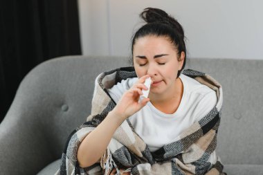 Kanepede oturan hasta genç bir kadının burun spreyi. Alerjik rhinitis semptomlar ve tedavi.