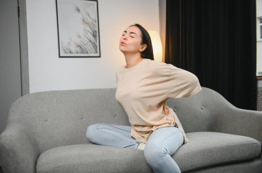 Sırt ağrısı çeken olgun bir kadın oturma odasındaki koltukta oturan mutsuz, yaşlı bir sarışın sırt ağrısı yüzünden rahatsız..