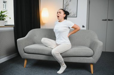 Sırt ağrısı çeken olgun bir kadın oturma odasındaki koltukta oturan mutsuz, yaşlı bir sarışın sırt ağrısı yüzünden rahatsız..