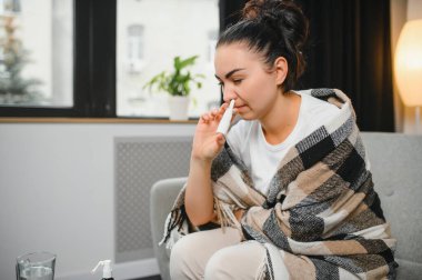 Kanepede oturan hasta genç bir kadının burun spreyi. Alerjik rhinitis semptomlar ve tedavi.
