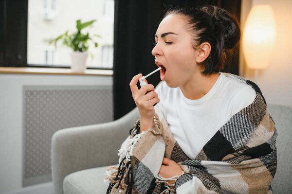 sick young woman in warm clothes spraying throat while sitting on couch at home