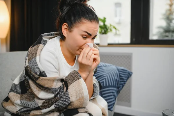 Ill young woman sit on sofa covered with blanket, freezing, blowing her runny nose, sneeze in tissue. Female got fever, flu and influenza symptoms cough at home. Sick allergic with respiratory disease.