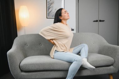 Sırt ağrısı çeken olgun bir kadın oturma odasındaki koltukta oturan mutsuz, yaşlı bir sarışın sırt ağrısı yüzünden rahatsız..