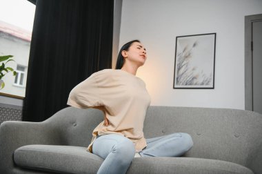 Sırt ağrısı çeken olgun bir kadın oturma odasındaki koltukta oturan mutsuz, yaşlı bir sarışın sırt ağrısı yüzünden rahatsız..