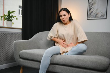 Genç bir kadın evdeki kanepede otururken karın ağrısı çekiyor. Karın ağrısının nedeni adet sancısı, mide iltihabı, mide ülseri, gıda zehirlenmesi, ishal ya da bağırsak iltihabı. Seçici odak.