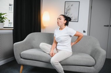 Sırt ağrısı çeken olgun bir kadın oturma odasındaki koltukta oturan mutsuz, yaşlı bir sarışın sırt ağrısı yüzünden rahatsız..