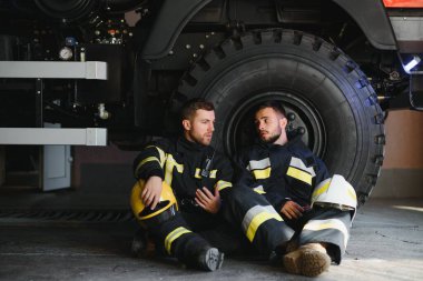 İtfaiye aracının önünde duran üniformalı ve kasklı iki profesyonel itfaiyeci.