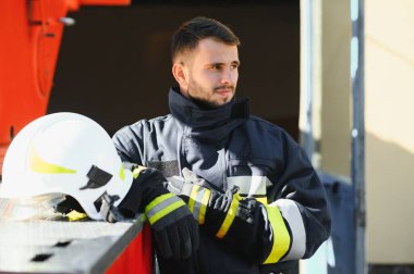 Görevdeyken itfaiyeci portresi. İtfaiye arabasının yanında gaz maskesi ve miğferi olan itfaiyeci.
