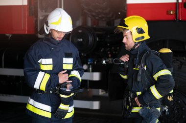 İtfaiye aracının önünde duran üniformalı ve kasklı iki profesyonel itfaiyeci.