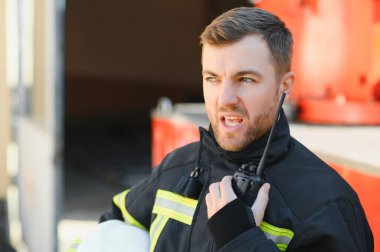 Koruyucu üniformalı itfaiyeci itfaiye aracının yanında duruyor ve telsizle konuşuyor.