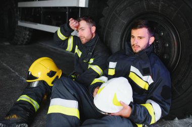 İtfaiye aracının önünde duran üniformalı ve kasklı iki profesyonel itfaiyeci.