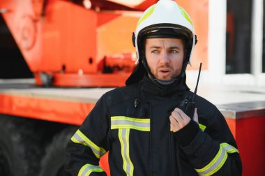 Koruyucu üniformalı itfaiyeci itfaiye aracının yanında duruyor ve telsizle konuşuyor.