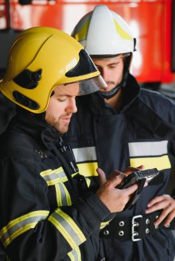 İtfaiye aracının önünde duran üniformalı ve kasklı iki profesyonel itfaiyeci.