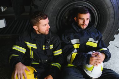 İtfaiye aracının önünde duran üniformalı ve kasklı iki profesyonel itfaiyeci.