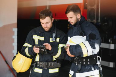 İtfaiye aracının önünde duran üniformalı ve kasklı iki profesyonel itfaiyeci.