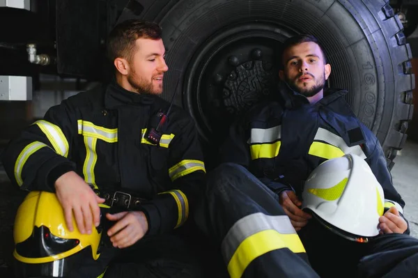 İtfaiye aracının önünde duran üniformalı ve kasklı iki profesyonel itfaiyeci.