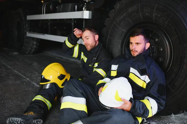 İtfaiye aracının önünde duran üniformalı ve kasklı iki profesyonel itfaiyeci.