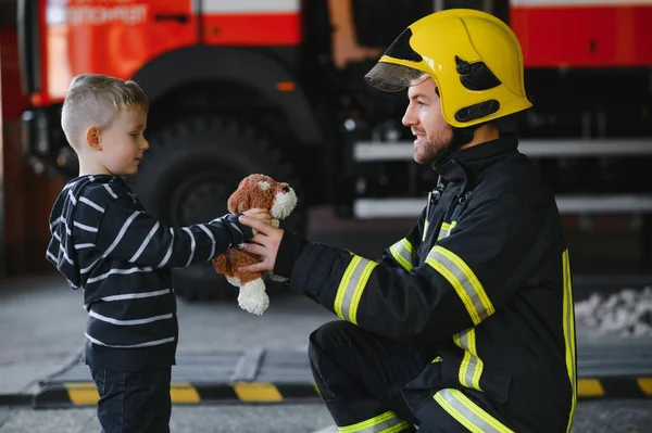 Dirty firefighter in uniform holding little saved boy standing on black background