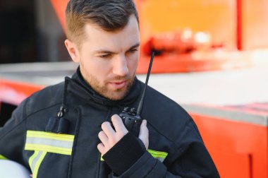 Koruyucu üniformalı itfaiyeci itfaiye aracının yanında duruyor ve telsizle konuşuyor.