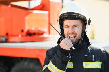 Koruyucu üniformalı itfaiyeci itfaiye aracının yanında duruyor ve telsizle konuşuyor.