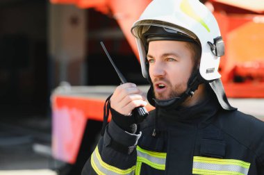 Koruyucu üniformalı itfaiyeci itfaiye aracının yanında duruyor ve telsizle konuşuyor.