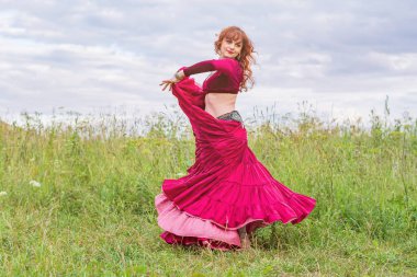 Kırmızı balo elbisesi giymiş güzel kızıl saçlı kız yeşil bir çayırda mavi gökyüzüne karşı ateşli bir dans ediyor..