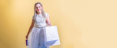 Saçları örgülü, beyaz elbiseli genç bir kız sarı arka planda duruyor ve elinde paketleri tutuyor. Kameraya bak..