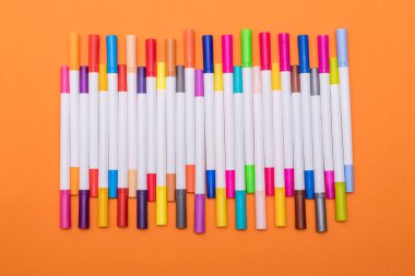 A set of multi-colored markers (felt-tip pens) lies in an even row on an orange background.