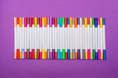 A set of multi-colored markers (felt-tip pens) lies in an even row on a purple background.