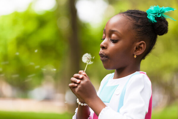 Outdoor portrait of a cute young black girl blowing a dandelion