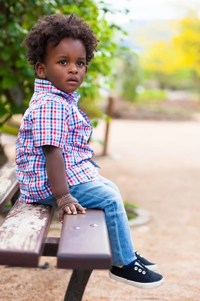 一个黑色的小男孩，坐落在一条长凳上的室外肖像 — 图库照片