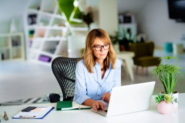 Kendine güveni tam orta yaşlı bir iş kadını laptopunun arkasında oturuyor ve yeni bir proje üzerinde çalışıyor.. 