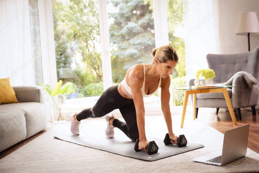 Shot of a woman using dumbbells while doing body workout at home. Woman wearing sports wear and using a laptop while exercising on the yoga mat. Healthy lifestyle.