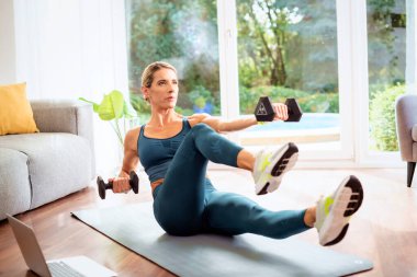 Evde vücut egzersizi yaparken bir kadının dambıl kullanması. Yoga minderinde egzersiz yaparken spor giyen ve dizüstü bilgisayar kullanan bir kadın. Sağlıklı yaşam tarzı.