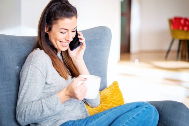 Neşeli kadın akıllı telefonunu kullanıyor ve evdeki koltukta dinlenirken telefonla konuşuyor.. 