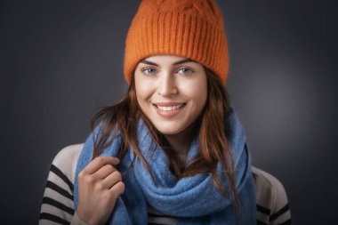 Kameraya bakıp gülümserken örgü şapka ve eşarp takmış genç bir kadının stüdyo fotoğrafı. Koyu arkaplanda izole. 