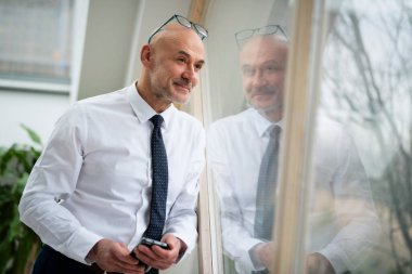 Gömlek ve kravat takmış gülümseyen bir işadamının pencerenin önünde dikilirken düşünceli bakışları..