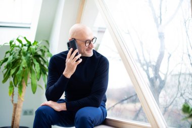 Balıkçı yaka kazak ve gözlük takan yakışıklı bir adamın pencerede otururken çekilmiş fotoğrafı.. 