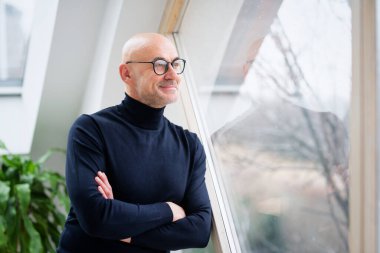 Kollarını katlayıp pencereden dışarı bakarken balıkçı yaka kazak giyen bir adam..