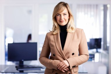 Ofiste dikilirken balıkçı yaka kazak ve ceket giyen güzel sarı saçlı iş kadını..